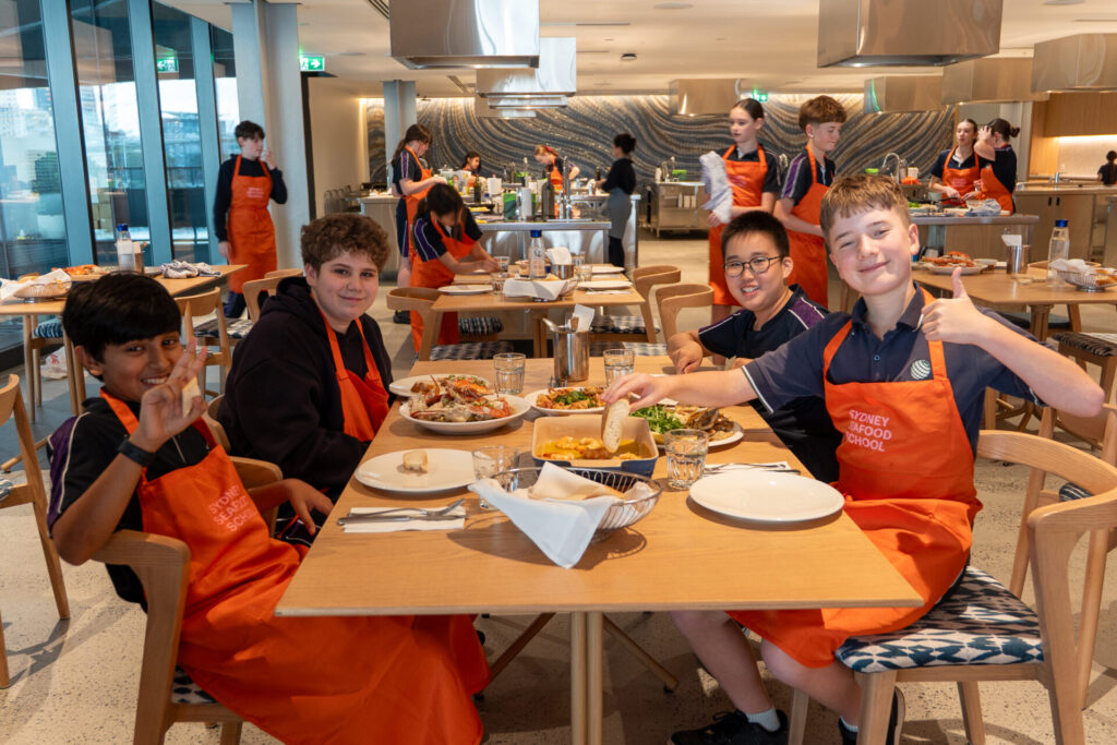 A group of secondary school students wearing orange aprons sit at a table in a modern kitchen classroom, smiling and giving thumbs up while enjoying a meal inspired by culinary fusion. Other students cook and work in the background.
