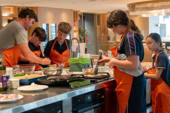 Five secondary school students in orange aprons participate in a cookery class, exploring culinary fusion as they prepare food together around a modern kitchen island at the Sydney Fish Market.