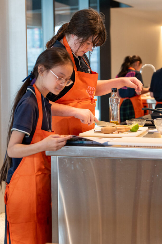 Two girls in orange aprons prepare food at a kitchen worktop, one cutting bread while the other checks a tablet—perhaps exploring a Culinary Fusion recipe. Various kitchen items are scattered across their secondary school cooking workspace.
