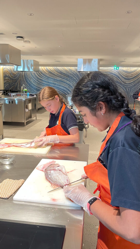 Two girls wearing orange aprons and gloves prepare fish at a stainless steel kitchen counter, focusing intently. The modern kitchen, reminiscent of Sydney Fish Market, features cooking stations and patterned walls in the background.