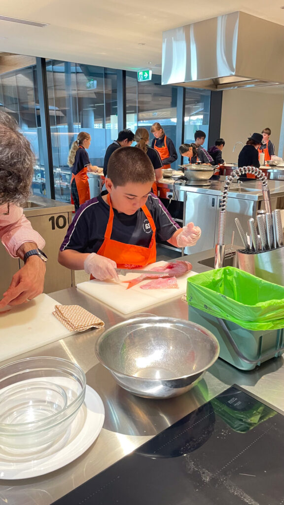 A secondary school boy wearing an orange apron and gloves prepares fish at a kitchen workstation, surrounded by bowls and utensils, as other children cook in the background—a scene inspired by the vibrant energy of Sydney Fish Market.