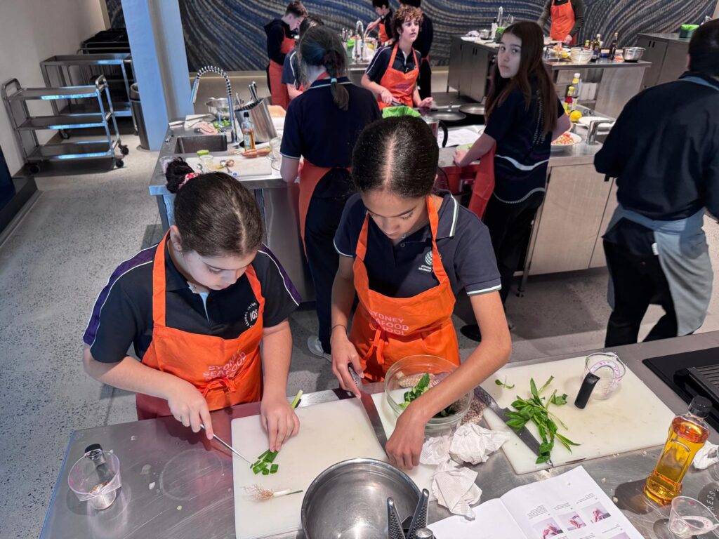 Two girls in orange aprons chop green vegetables at a kitchen workstation during a secondary school culinary fusion class, while other students in similar aprons cook and prepare food in the background of a modern kitchen classroom.