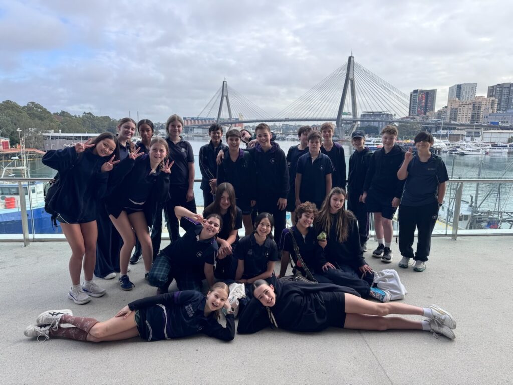 A group of secondary school students in dark uniforms pose for a photo on a waterfront terrace, with a large bridge, boats, and city buildings—possibly near the Sydney Fish Market—in the background; four students lie at the front, smiling.