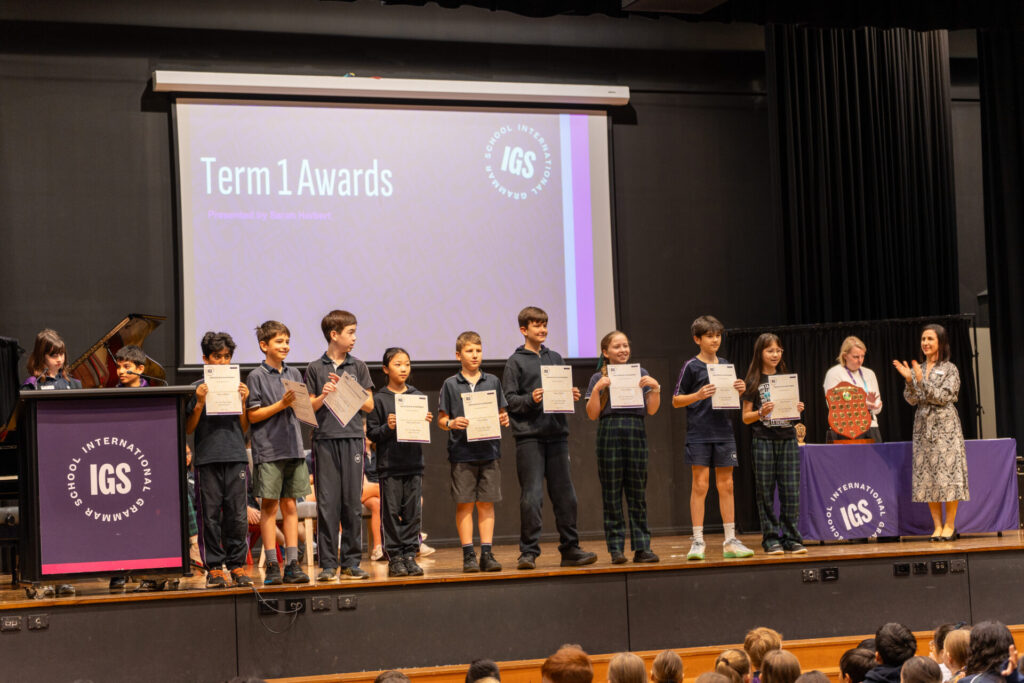 A group of students stands onstage holding certificates during a Term 1 Awards ceremony celebrating sporting achievements. A woman stands at a lectern, while others, including an adult, applaud from the side. An audience watches from below.