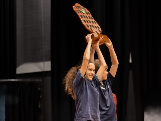 Two children in navy shirts smile and lift a large wooden shield-shaped trophy above their heads on stage, celebrating their term sports achievements against a backdrop of black curtains.