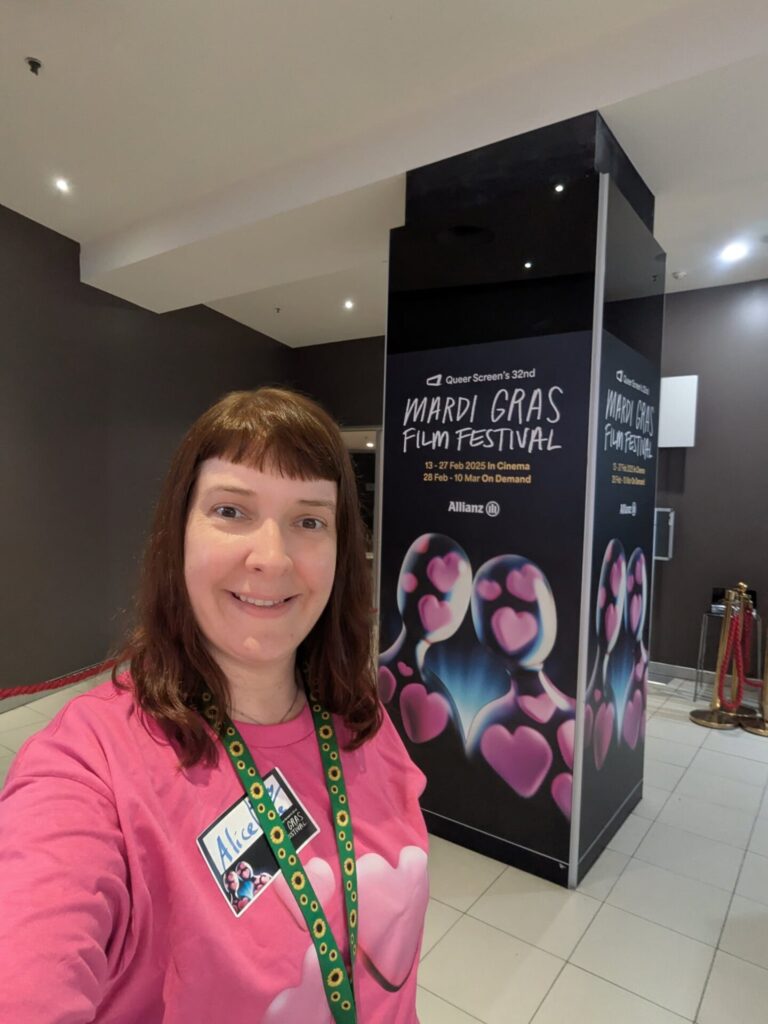 A smiling person with straight brown hair and a pink shirt stands indoors near a large display for the Mardi Gras Film Festival, decorated with heart and film reel graphics.