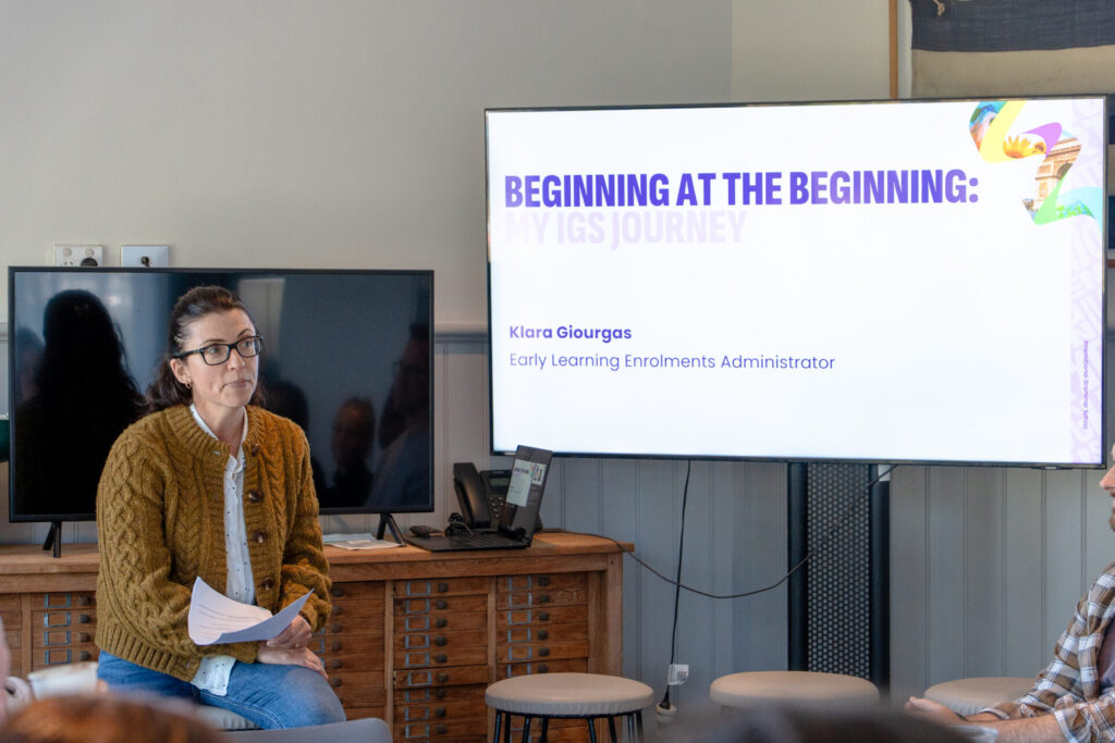 A woman wearing glasses and a brown jumper sits holding papers next to a screen displaying a presentation titled BEGINNING AT THE BEGINNING: KLARA GIOURGAS, Early Learning Enrolments Administrator.
