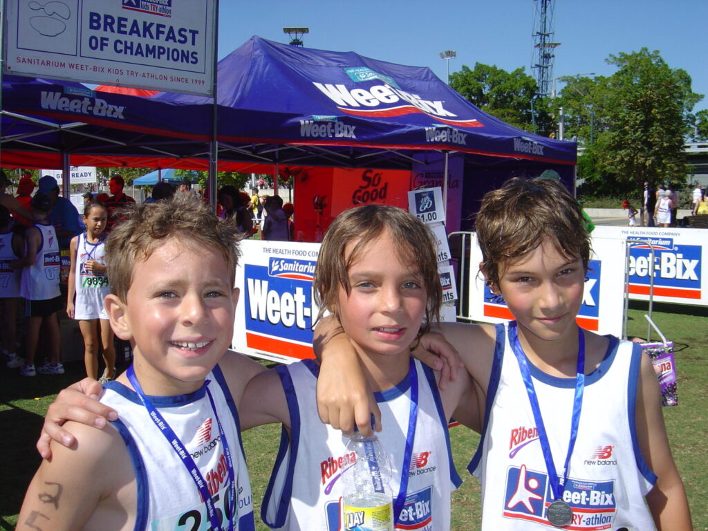 Three smiling children wearing sports kit and medals stand together with arms around each other at an outdoor sports event, in front of a Weetabix tent. Other participants and green trees are visible in the background.