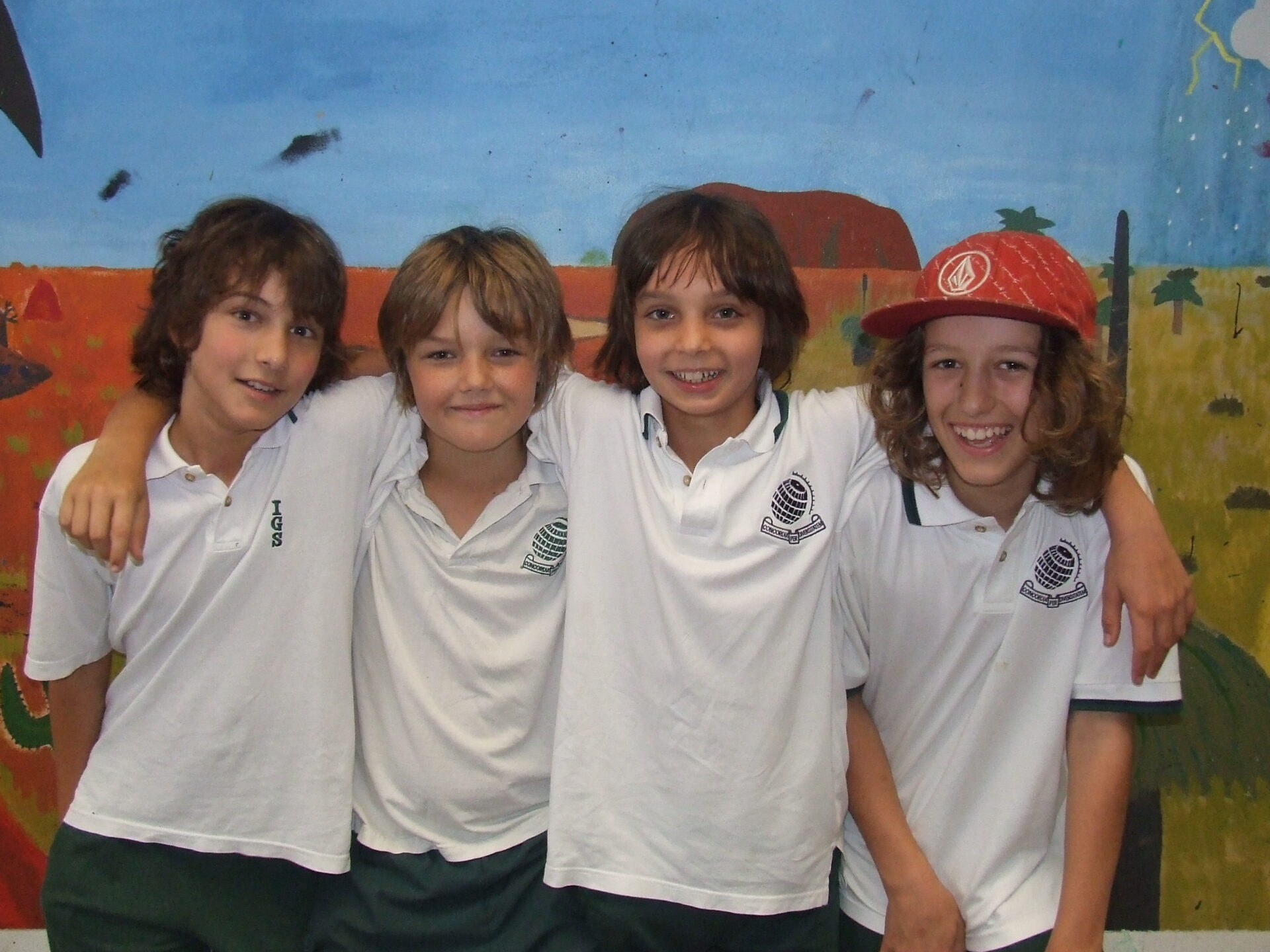 Four smiling children in white school uniforms stand close together in front of a colourful mural, with three of them linking arms. One child wears a red cap.