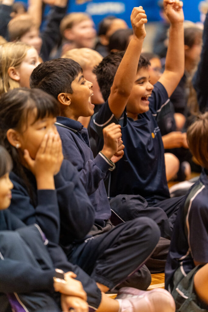 A group of schoolchildren sitting on the floor cheer and celebrate excitedly, some with hands raised and wide smiles, while one girl covers her mouth in surprise. Their faces reflect the Sound of Creativity as they watch an event or performance unfold.