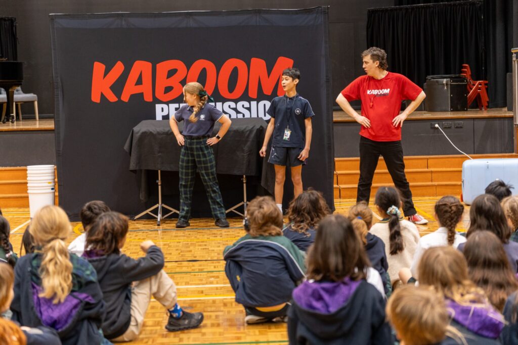 Three people stand on stage in front of a Kaboom Percussion banner, bringing the Sound of Creativity to life for an audience of seated children in a gym or auditorium. Two wear navy outfits, and one sports a red Kaboom shirt.
