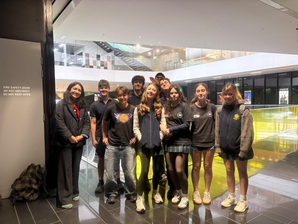 A group of nine students in casual and school uniforms stand together indoors, smiling, with a woman in business attire on the left. They gather in a modern building with glass balustrades and bright lighting, reflecting a shared culture and connection through language.