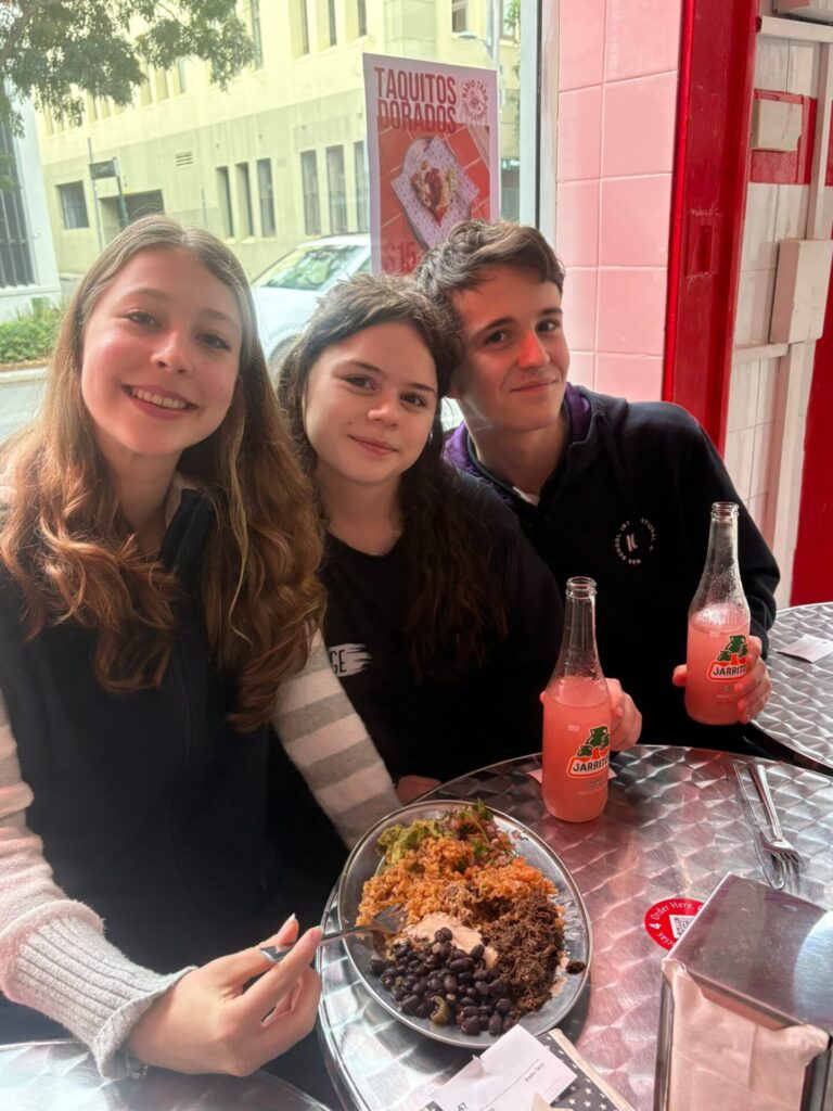 Three smiling young people sit at a metal table inside a restaurant, sharing Mexican food and Jarritos fizzy drink. A Taquitos Dorados poster in the background highlights the rich culture visible through the window behind them.