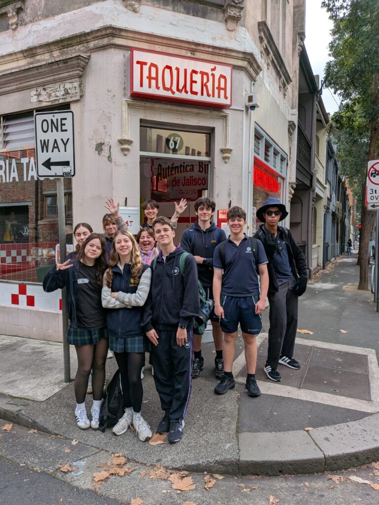 A group of nine smiling teenagers in school uniforms stand on a city street corner in front of a taqueria, offering a window into language and culture as they wave and pose for the photo. A One Way street sign is visible nearby.