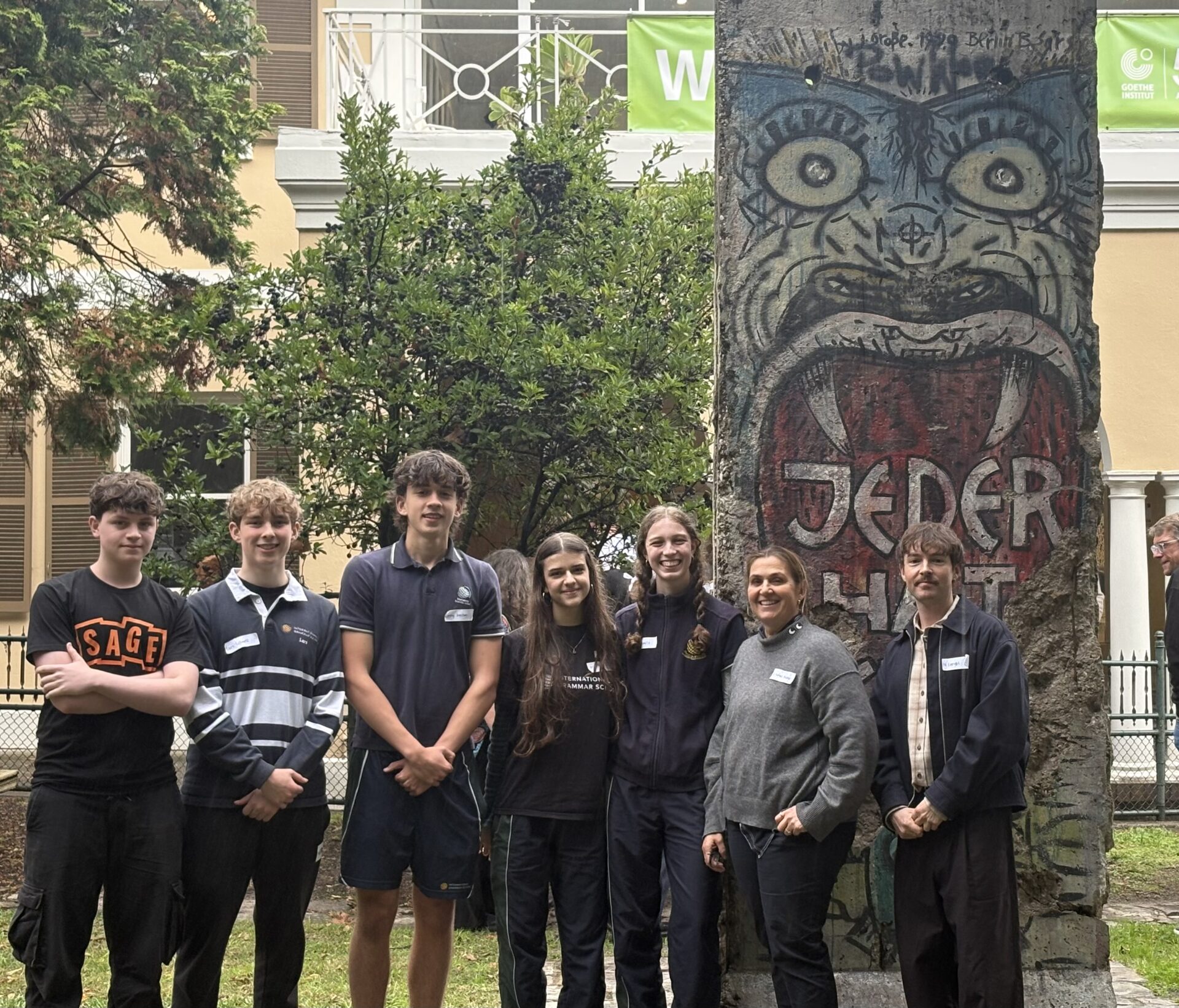 Seven people stand smiling in front of a painted piece of the Berlin Wall with JEDER HUT written on it, outside a building with windows, trees, and greenery—a scene capturing culture and connection.