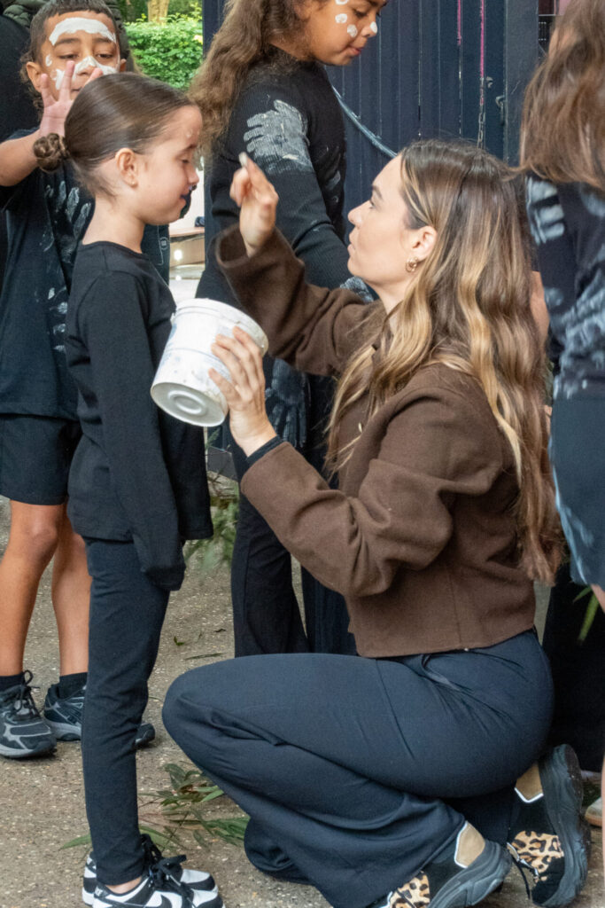 A woman kneels to paint a young girl's face at an IGS NAIDOC Day celebration, holding a container whilst surrounded by other children with face paint outdoors. The children and woman are dressed mostly in black.