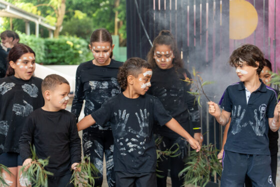 A group of children from IGS, wearing black clothes with white handprint designs and face paint, stand together holding green branches, appearing to participate in a cultural or traditional NAIDOC Day activity.