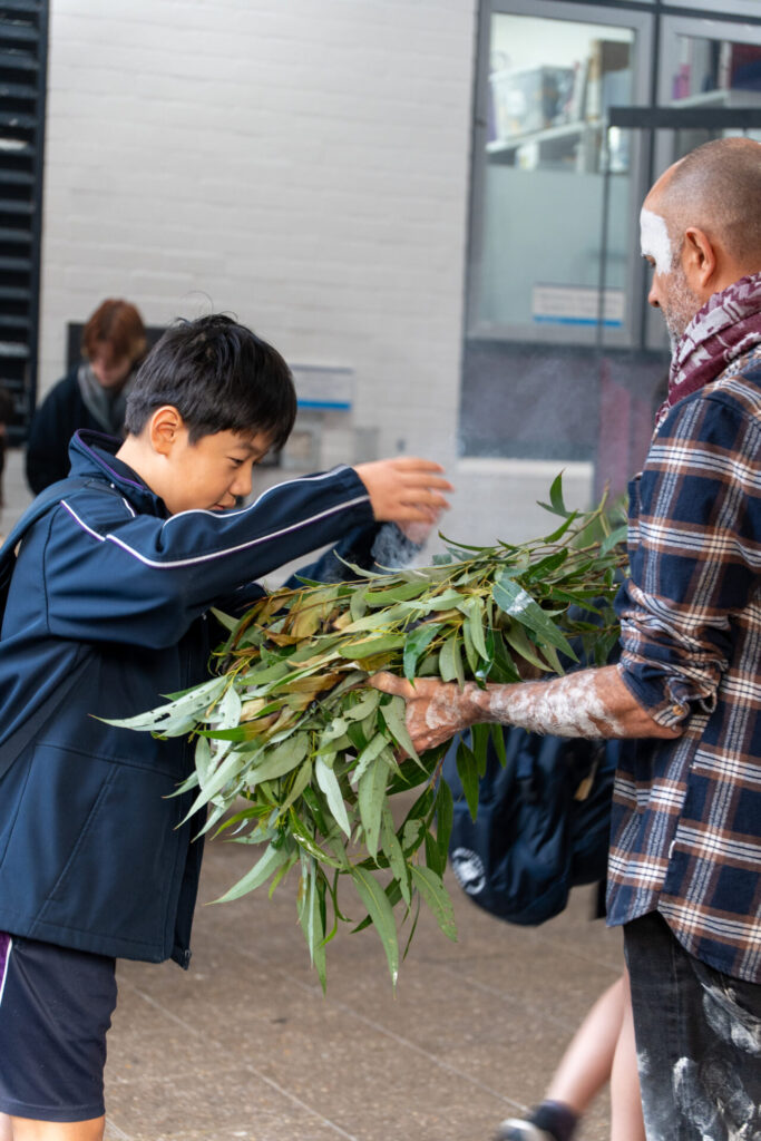 A boy in a navy jacket participates in a NAIDOC Day ceremony, wafting smoke from eucalyptus leaves held by an adult with painted face and arm, in an indoor setting at IGS.