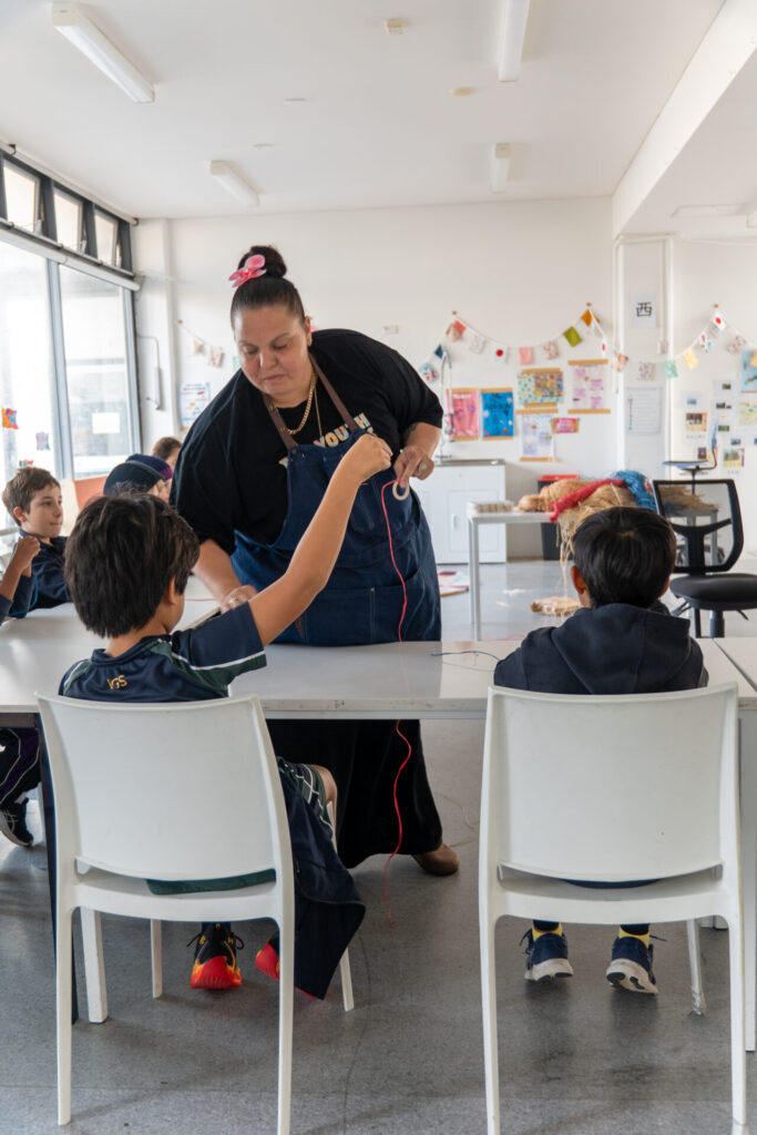 A teacher in a blue apron leans over a table to help a young pupil with string in a bright classroom during an IGS NAIDOC celebration, whilst other children sit and work on their own activities.