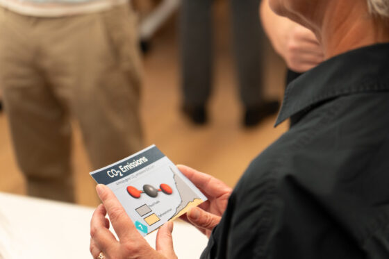 A person holds a card labelled CO₂ Emissions with a chart and coloured ovals, highlighting Eco-School research. Two others stand nearby, but the focus remains on the card and the person’s hands.