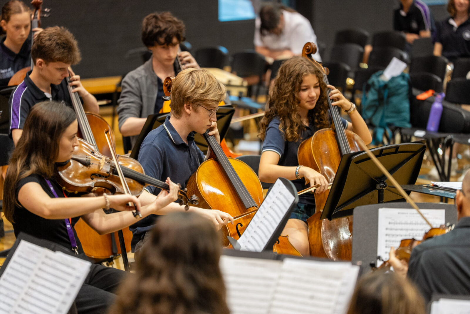 A group of young musicians play string instruments, including violins and cellos, whilst reading sheet music during a rehearsal in a concert hall, showcasing the results of dedicated music tuition and collaborative practice in music ensembles.