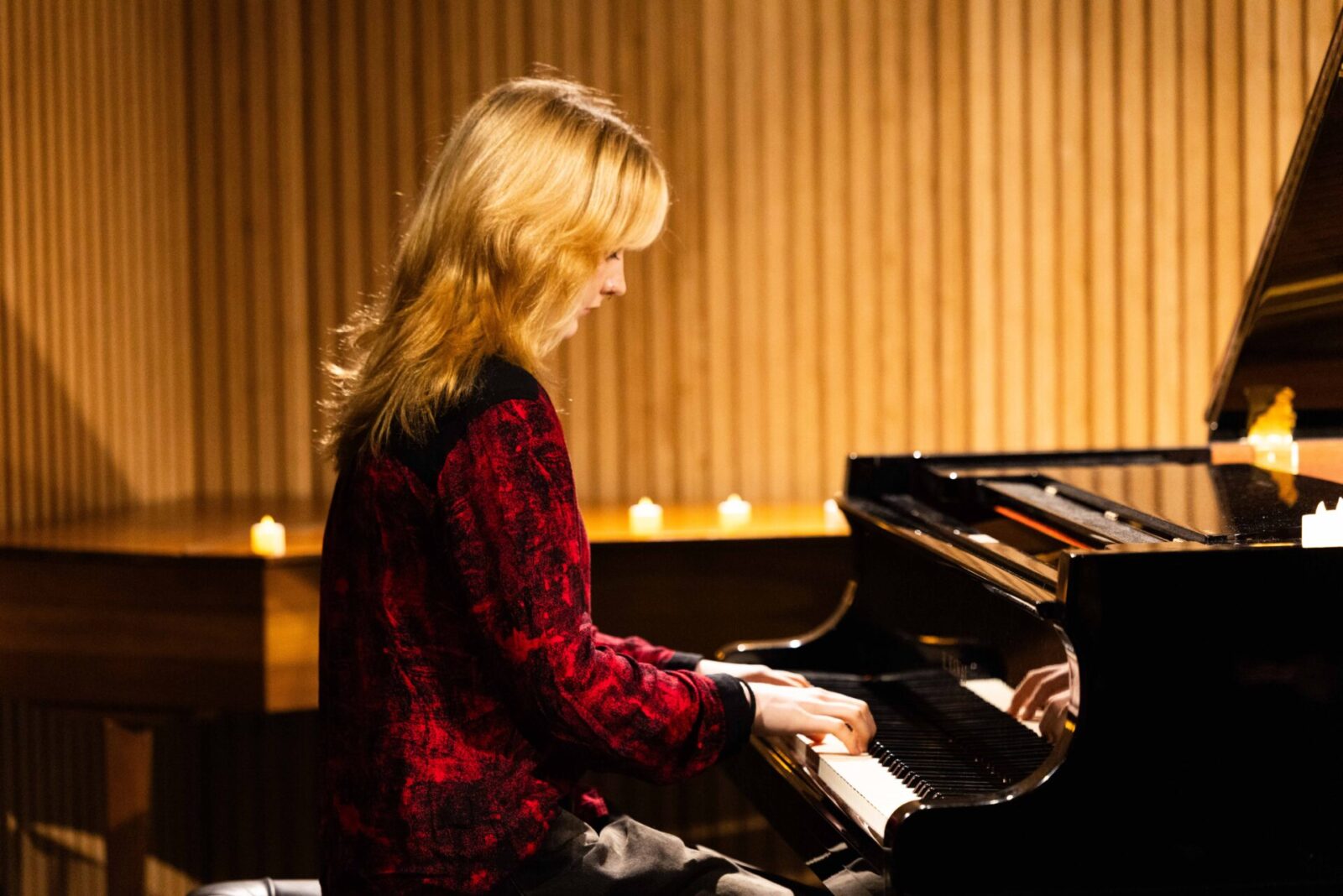A person with fair hair wearing a red and black jacket plays a grand piano in a warmly lit room, where candles rest on the piano and wood-panelled walls surround the scene—a captivating setting often found in music ensembles or during personalised music tuition sessions.