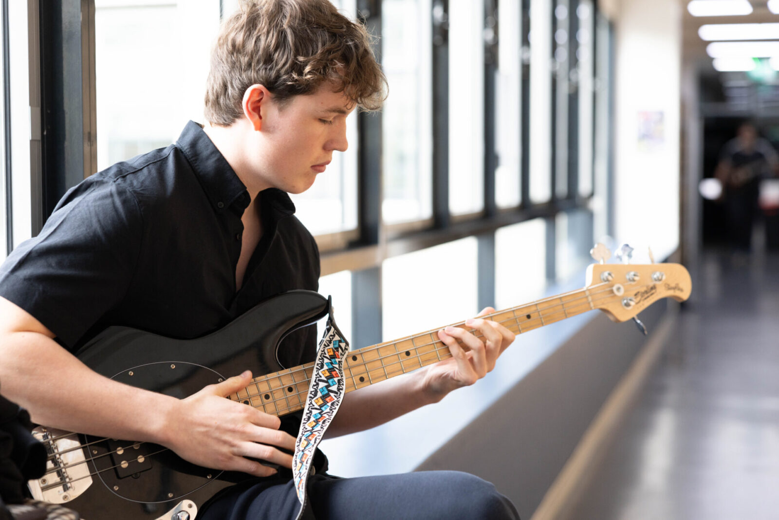 A young man in a black shirt sits by large windows, playing a black electric bass guitar with a colourful strap, focused on his music tuition in a bright corridor.