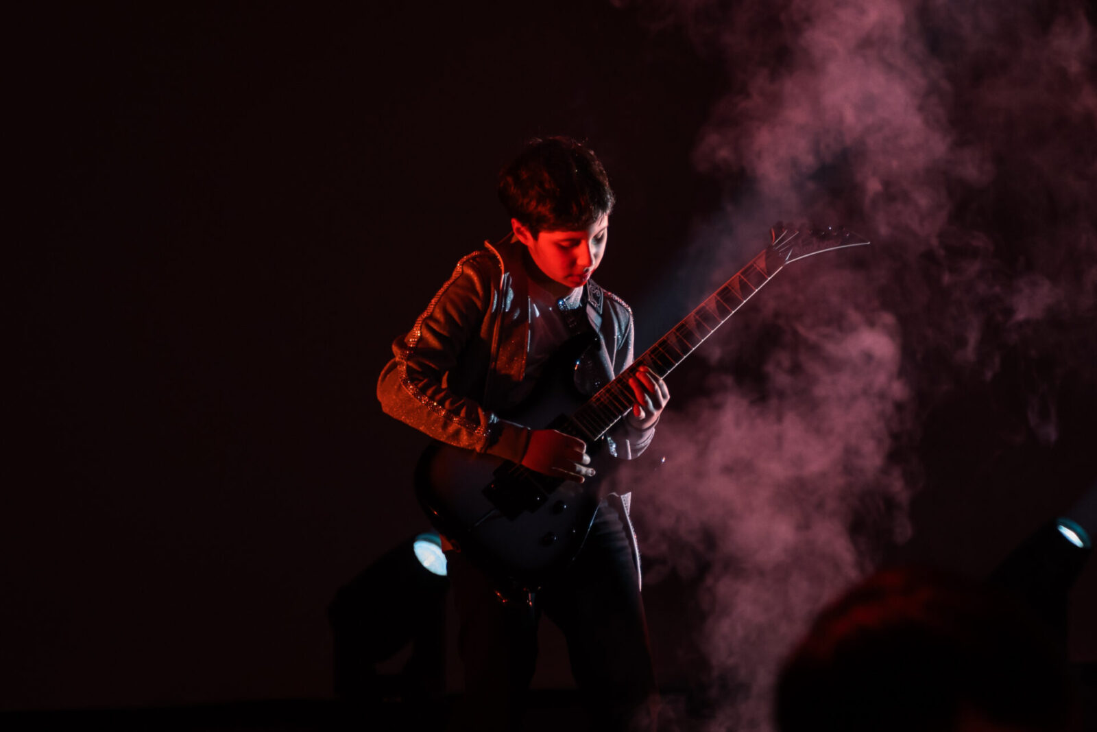 A young boy plays an electric guitar on stage, surrounded by dramatic red lighting and smoke effects, captivating the audience as he performs with IGS ensembles.