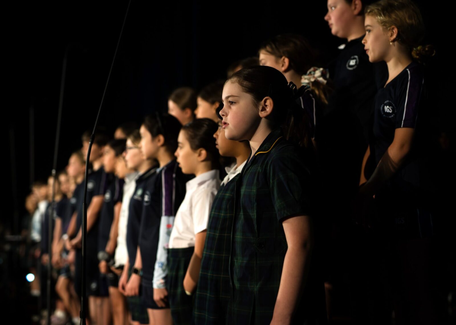 A group of children wearing school uniforms stand in a queue on stage, facing forwards, appearing to sing or perform as part of one of the school's music ensembles in a darkened auditorium.