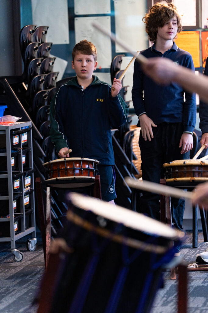 Two boys stand in a classroom playing taiko drums. One, appearing focused, holds drumsticks while the other looks on, both participating in a lively Day of Taiko. Blurred drumsticks and drums are visible in the foreground, with stacked chairs and shelves in the background—capturing a vibrant Year 6 music lesson.