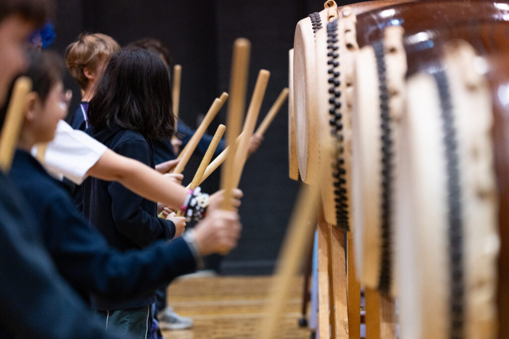 Children in Year 6 stand in a queue playing traditional Japanese taiko drums with wooden sticks, focusing intently on their performance during the Day of Taiko.