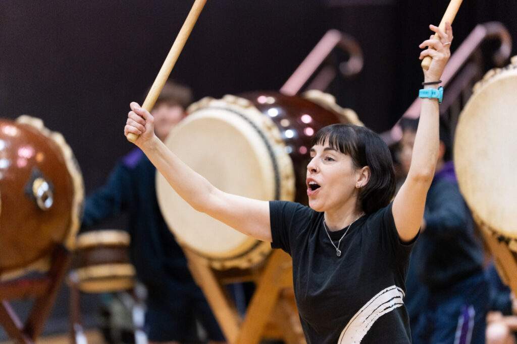 A person enthusiastically plays taiko drums, holding drumsticks above their head with both hands, as part of the vibrant Day of Taiko celebration. Large drums and other participants—including Year 6 pupils—are visible in the background.