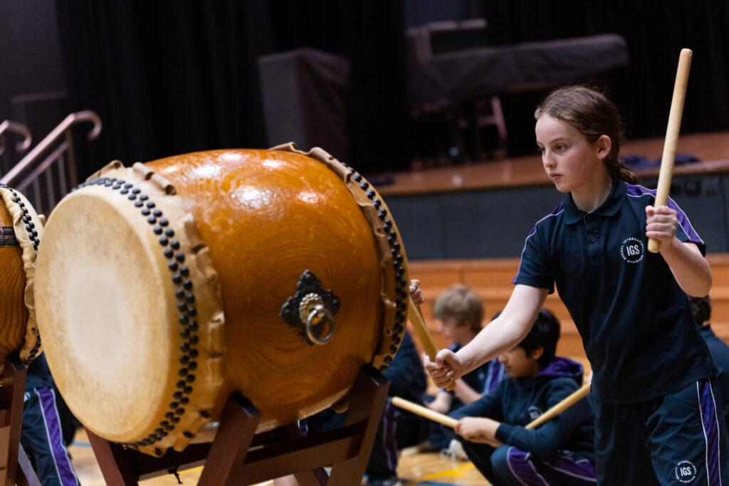 A Year 6 girl in a dark uniform plays a large traditional Taiko drum with drumsticks in a gymnasium while others watch or wait their turn in the background, celebrating the Day of Taiko.