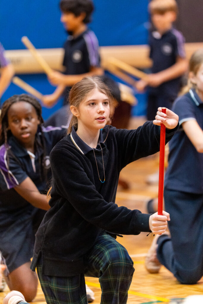 A Year 6 girl kneels indoors, holding a red stick, with other children participating in a Day of Taiko activity in the background. All wear school uniforms.