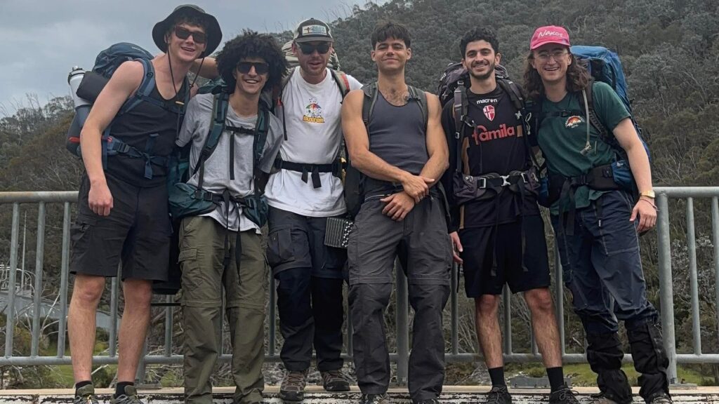 Six young adults in hiking gear stand close together and smile on a bridge, with forested hills and cloudy skies in the background, proudly displaying their TRIPLEPLAY badges after completing number 36 on the WITWAT trail list.