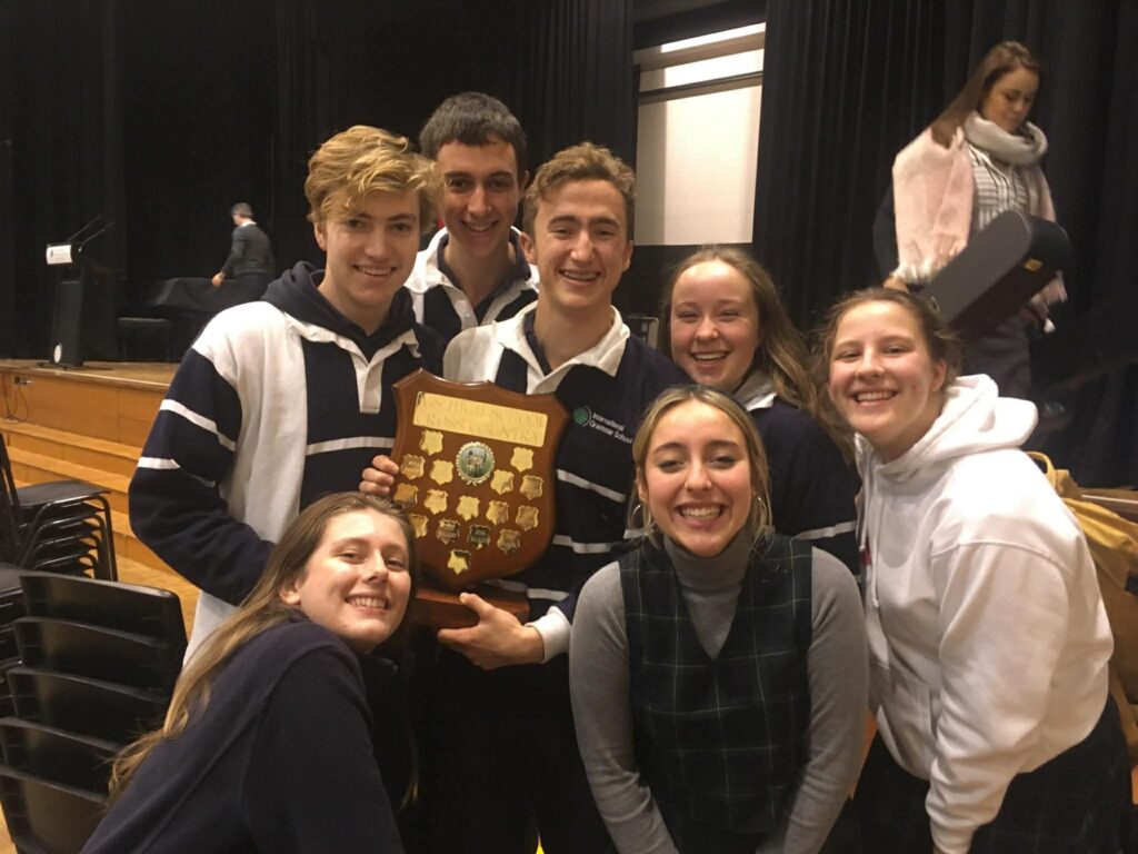 Six smiling young people pose indoors, holding a wooden shield-shaped trophy with gold plates. They wear casual clothes and stand in front of a stage with black curtains, proudly celebrating their achievement at the WITWAT TRIPLEPLAY #36 event.