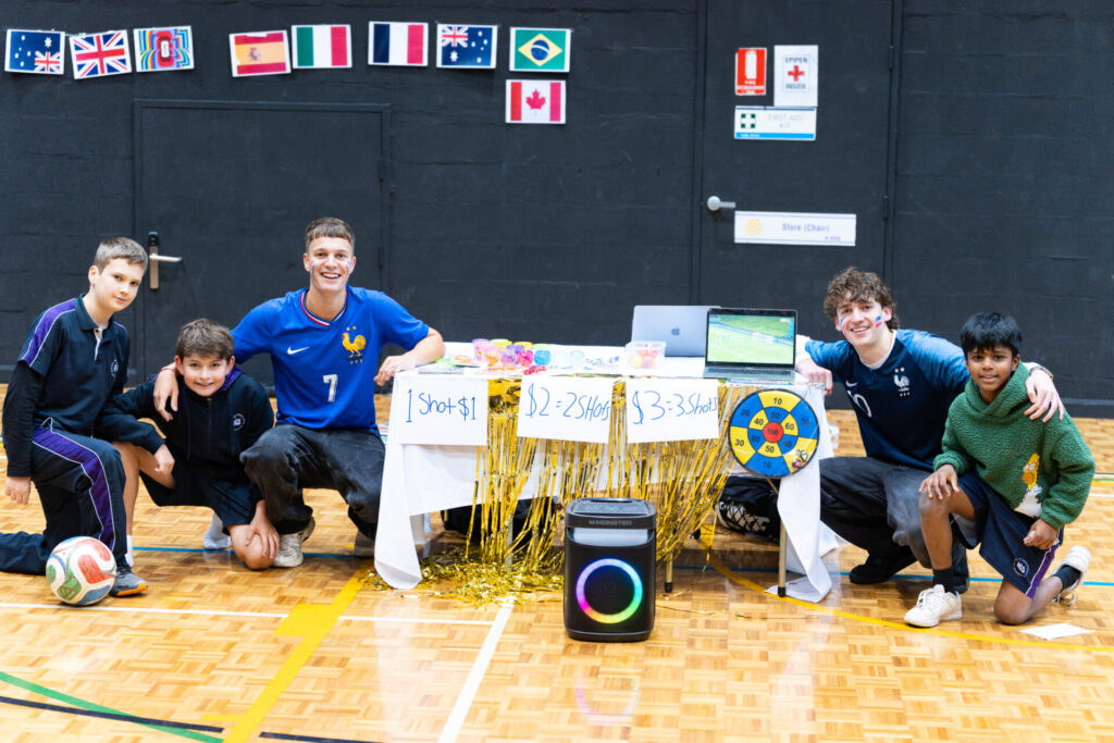Five boys kneel and smile by a decorated football-themed stall in a gym, with flags and a football visible. Showcasing the spirit of tomorrow’s entrepreneurs, the group has set up a laptop displaying a game and arranged several prizes on the table, reflecting their enthusiasm for enterprise.