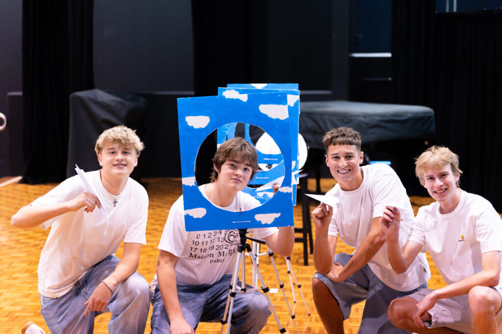 Four smiling teenagers in white shirts kneel indoors, each holding a paper aeroplane. They pose with a blue board decorated with clouds and a circular cut-out, suggesting an engaging enterprise activity or event for future business leaders.