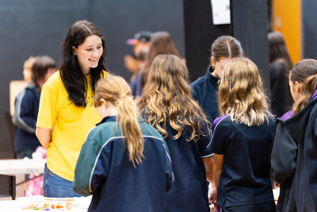 A woman in a yellow shirt smiles whilst talking to a group of children—Tomorrow's Entrepreneurs—gathered round a table with crafts or snacks, indoors at a busy event.