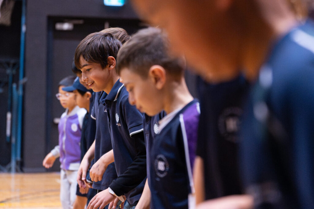 A group of children in sports kit stand in a row indoors, focused and ready, as if preparing to start a race or activity.