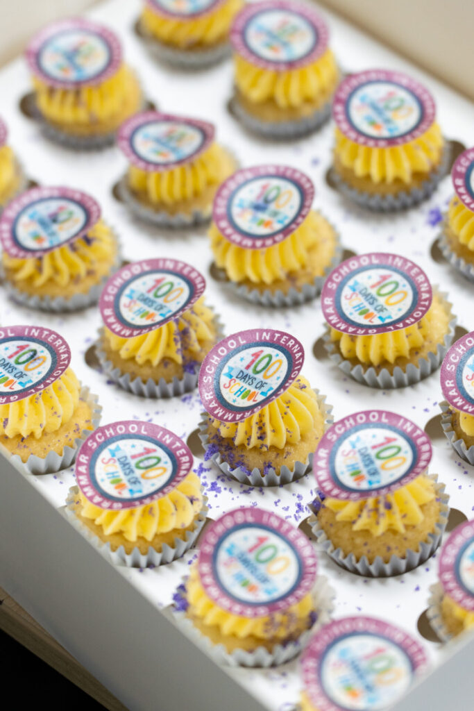 Mini fairy cakes with yellow icing, each topped with a round edible logo reading “100 Years” and “School of International Affairs”, arranged neatly in a white box.