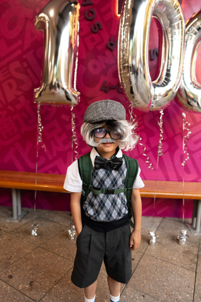 A young child dressed as an elderly person stands in front of large silver 100 balloons, wearing a grey wig, glasses, a bow tie, and a green rucksack, playfully capturing the spirit of 100 Days of Growth against a bright pink background.