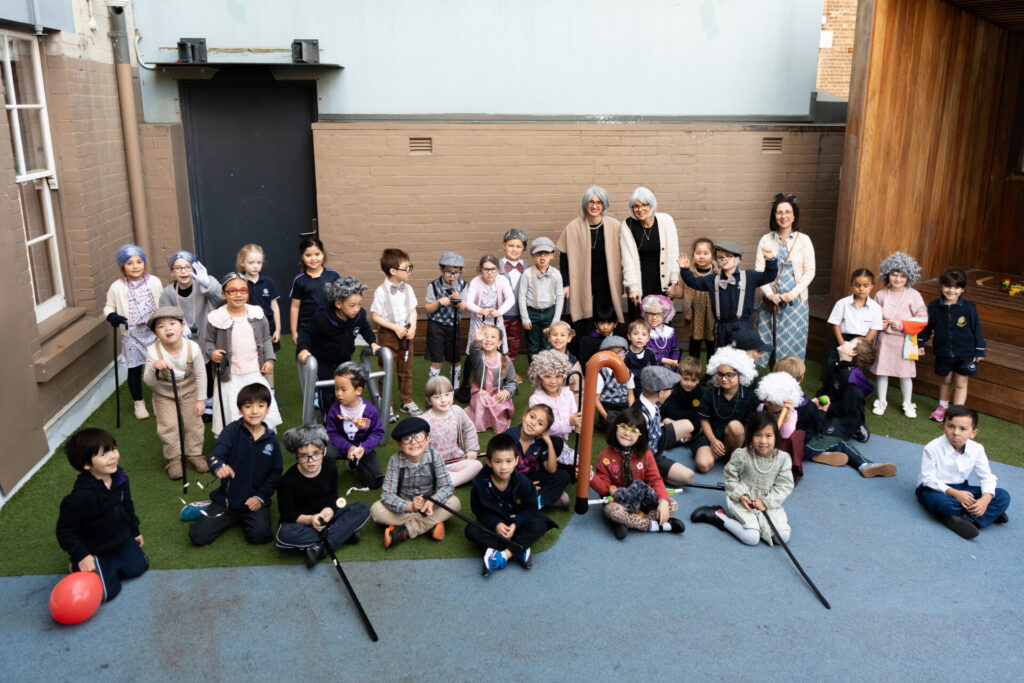 A group of young children and four adults pose outside, celebrating 100 Days of Growth. Many kids wear fancy dress, wigs, and glasses, resembling elderly people. They sit or stand on artificial grass and blue flooring, smiling at the camera.