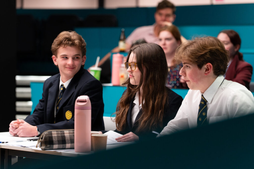 Three students in school uniforms sit at a desk in a classroom, engaging with each other whilst others sit in the background. A pink water bottle and checked pencil case are on the desk.