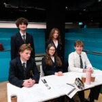 Five students in school uniforms sit and stand around a table with papers, pens, drinks, and pencil cases in a modern classroom with teal dividers. They appear to be taking part in a group activity or discussion.