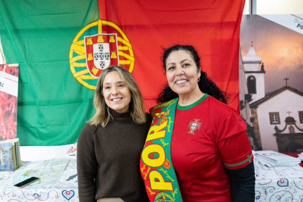 Two women smiling in front of a large Portuguese flag; one wears a brown jumper, the other a red Portugal shirt and scarf. It’s where all stories begin—at a stall with Portuguese-themed items and a poster of a church, proudly celebrating their country in the background.