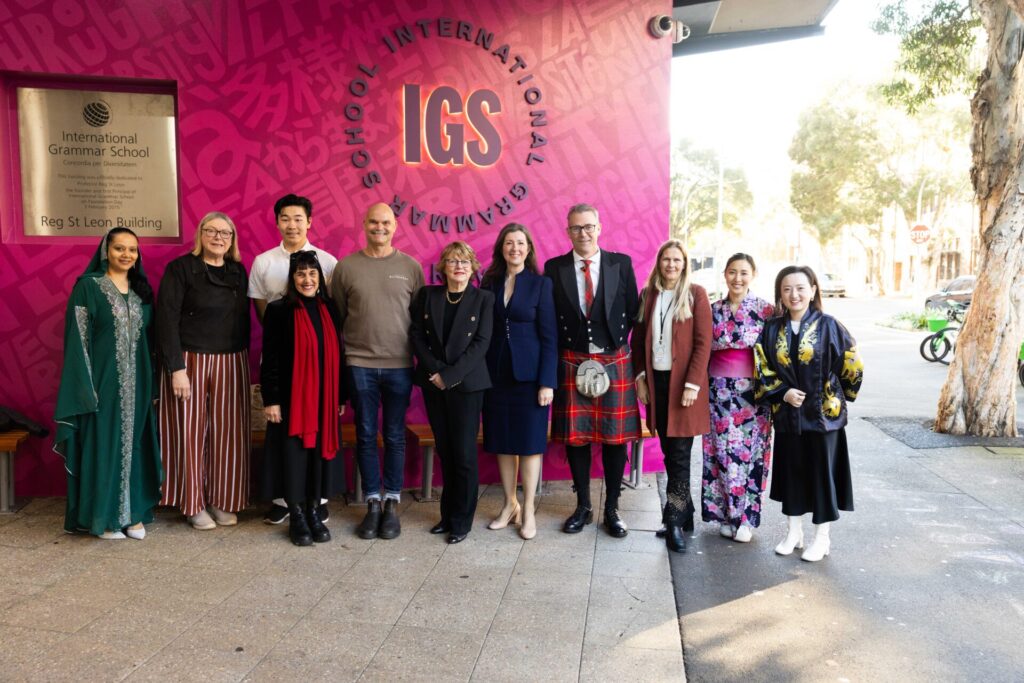 A diverse group of people stands smiling in front of a bright pink wall with the IGS logo and a school sign, where stories begin. They wear a mix of formal, casual, and traditional clothing, representing many countries—showing that at IGS, all stories matter.