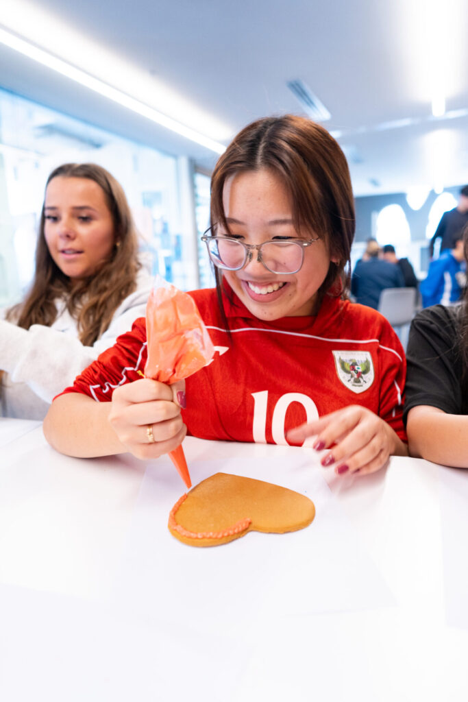 A smiling person in glasses decorates a heart-shaped biscuit with orange icing while sitting at a table, sharing sweet moments and stories with friends. Another person sits nearby, and others are visible in the background, creating a warm and lively atmosphere that feels right at home in any country.