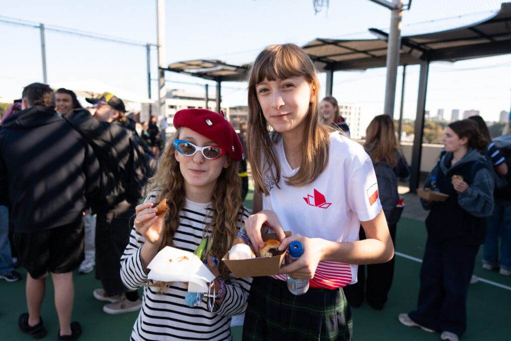 Two girls stand outdoors at an event, holding food trays. One wears a red beret and sunglasses, while the other sports a white shirt with a red logo. Amidst the lively crowd and bustling cityscape in the background, their presence hints at stories unfolding in this vibrant corner of the country.