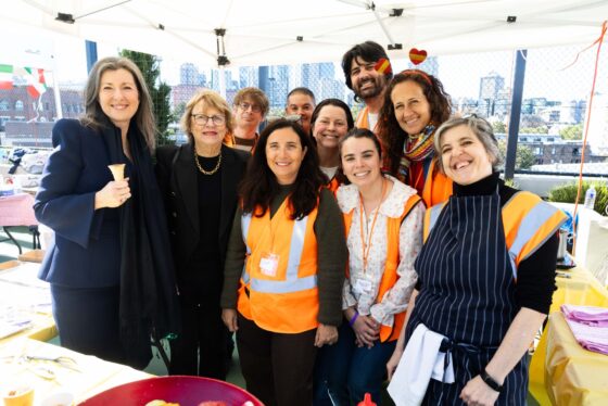 A group of ten people, some in orange safety vests, smile together under a marquee at an outdoor event with city buildings in the background. Embodying the spirit of Term in Full Flight, one woman holds an ice cream cone.